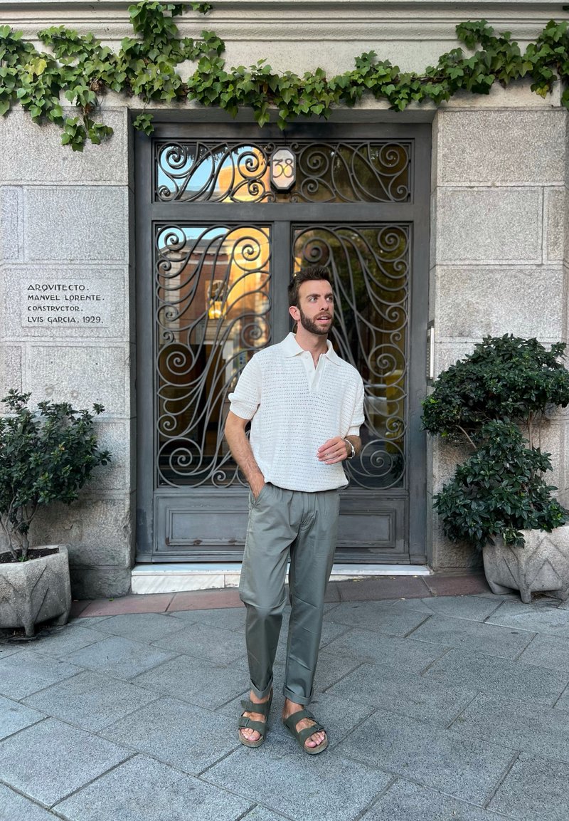 Αναπαυτική εμφάνιση που περιλαμβάνει ένα λευκό, υφασμάτινο πουκάμισο πόλο, ελιές παντελόνια και καφέ σανδάλια, σε φόντο από μια περίτεχνη μεταλλική πύλη και πράσινα φυτά.