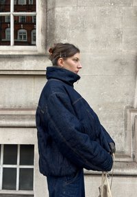 Tumša džinsa pūkains jaka ar augstu apkakli, izteiktiem šuvumiem un brīvu piegriezumu. Audums ir gluds, kas papildina tumšās bikses.