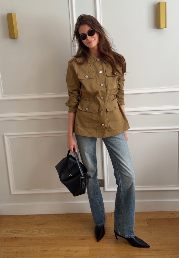 Vrouw draagt een beige jasje met knopen, blauwe jeans, zwarte puntige hakken, zonnebril en houdt een zwarte handtas vast, staand binnen tegen een witte muur.