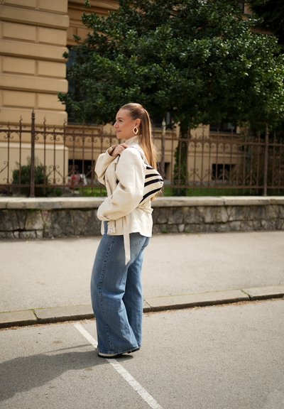 Joven mujer de pie en la calle con chaqueta beige, vaqueros de pierna ancha, mochila a rayas y pendientes geométricos con árboles y barandilla detrás.