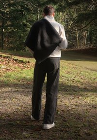 Άτομο με λευκό μπουφάν, μαύρο παντελόνι και λευκά παπούτσια, που φοράει μια μαύρη κουβέρτα στον ώμο, στέκεται σε ένα φωτεινό μονοπάτι δάσους.