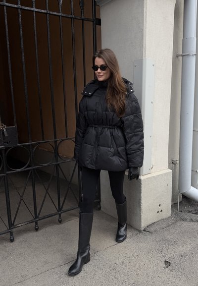 Mujer con chaqueta acolchada negra, leggings, botas hasta la rodilla, guantes y gafas de sol caminando cerca de una puerta de metal negra y una pared de edificio beige.