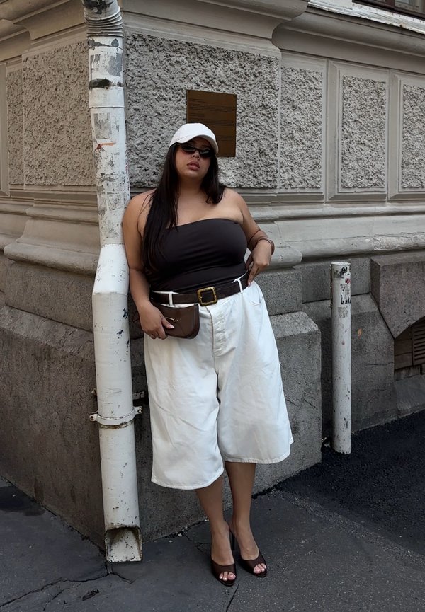 Femme portant une casquette blanche, un top bustier noir, un short blanc large et des talons noirs, appuyée contre un mur de pierre texturé et un tuyau blanc.