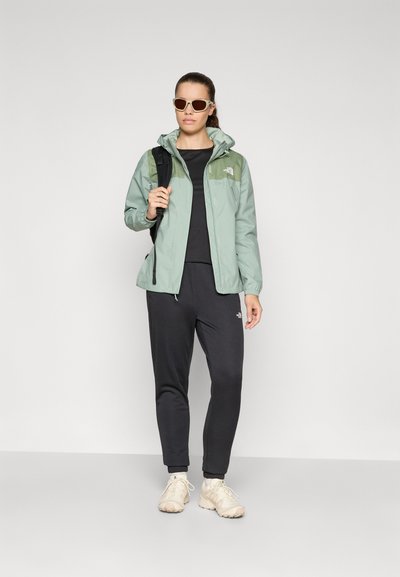 Chaqueta verde claro con capucha, camiseta negra y pantalones oscuros. Zapatos beiges y gafas de sol. Textura suave con un diseño deportivo.