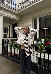 Maglia croppata beige chiaro con un bottone, abbinata a una sciarpa a righe nera e crema e pantaloni larghi scuri, ambientata su uno sfondo di balcone.