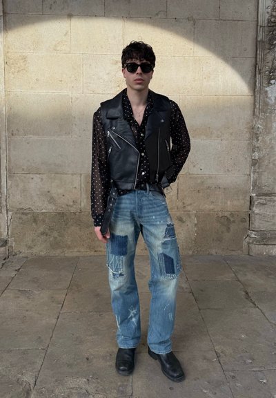Chaleco negro de cuero sin mangas sobre una camisa negra traslúcida con lunares, combinado con unos vaqueros azul desgastados con parches. Botines negros.