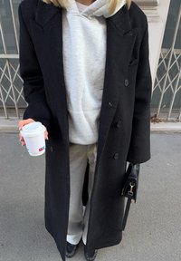 Personne portant un long manteau noir par-dessus un sweat à capuche gris et un pantalon, tenant un gobelet de café à emporter blanc et un sac à main noir sur un trottoir en ville.