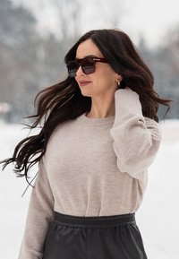 Femme aux longs cheveux foncés portant de grandes lunettes de soleil, un pull beige et un pantalon noir, debout en plein air dans un décor enneigé, la main touchant ses cheveux.