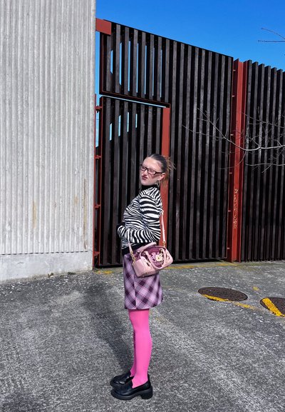 Mujer con top a rayas de cebra, falda de cuadros púrpura, medias rosas brillantes y zapatos negros, de pie de lado junto a una valla alta de madera negra y roja.