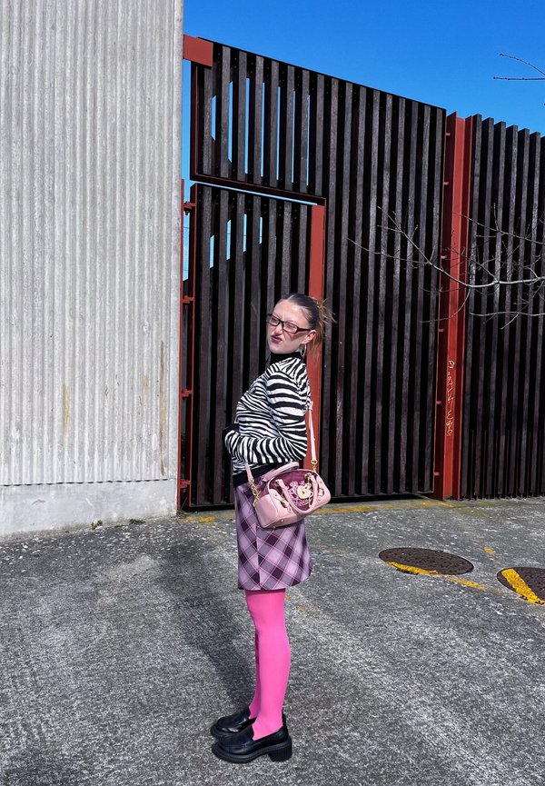 Femme en haut rayé zèbre, jupe à carreaux violette, collants rose vif et chaussures noires, debout de côté près d'une haute clôture en bois noire et rouge.