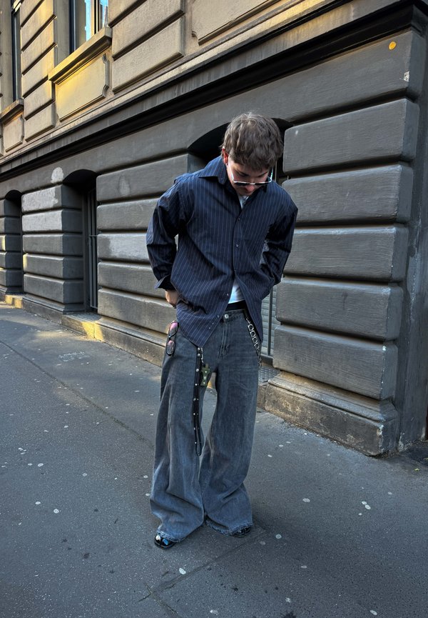 Camisa de manga larga a rayas negras, pantalones de mezclilla gris holgados, cadenas en la cintura y zapatos negros brillantes son elementos destacados.