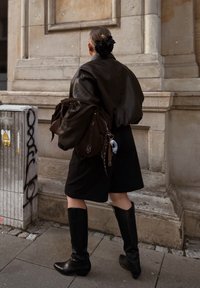 Žena kráčí poblíž kamenné zdi, na sobě má černou koženou bundu, sukně až ke kolenům, vysoké černé boty a nese velkou tmavě hnědou tašku, vlasy má sepnuté černým gumičkou.