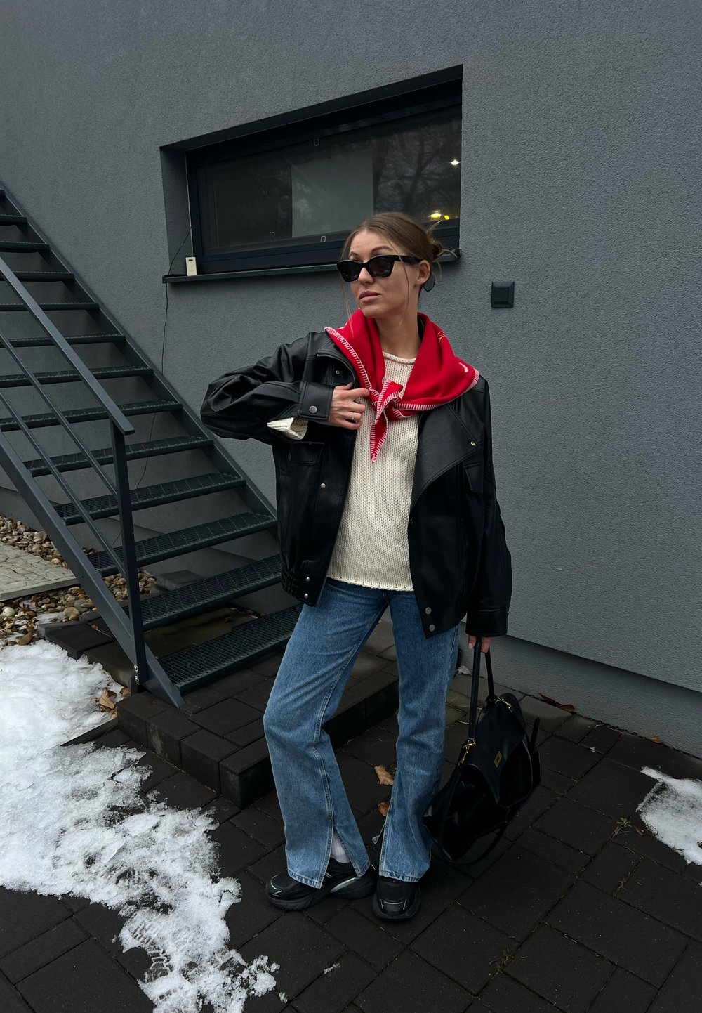 Femme portant des lunettes de soleil, une veste en cuir noire, une écharpe rouge, un pull blanc, un jean bleu et des chaussures noires, debout près d'un escalier métallique et de plaques de neige.