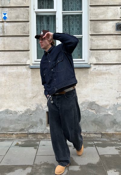 Chaqueta de mezclilla con dos bolsillos en el pecho, de color azul oscuro; pantalones negros holgados, botas marrones y una gorra marrón. De pie en una calle de adoquines.