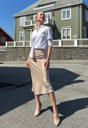 Camisa blanca de botones, falda midi de satén beige y tacones con tira al tobillo brillantes. Textura suave, diseño ajustado y paleta de colores neutros.