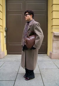 Borsa a mano in pelle marrone con forma arrotondata. La persona indossa un lungo cappotto marrone, una sciarpa nera con motivo e pantaloni scuri con scarpe color amaranto.