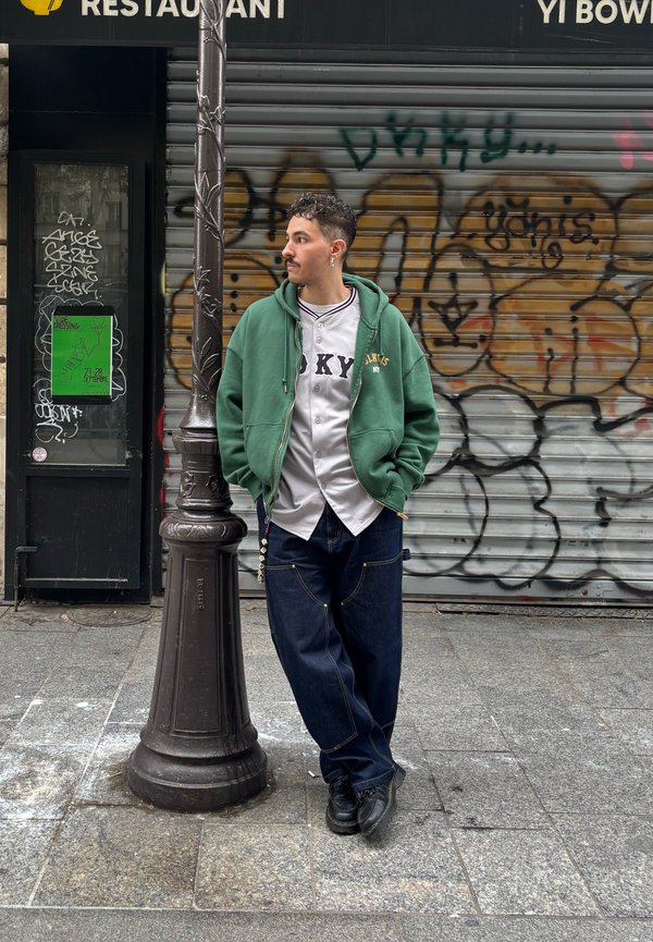 Sweatshirt verde sobre uma camisola branca, jeans largos azuis e sapatos pretos. Em pé ao lado de um poste de luz com graffiti e um cartaz verde ao fundo.