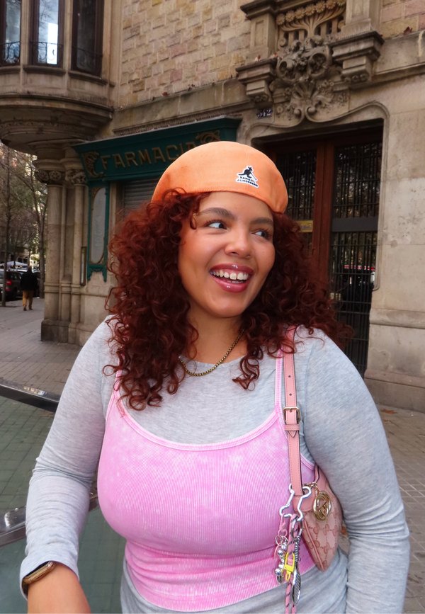 Jonge vrouw met krullend rood haar draagt een oranje pet, een roze top over een grijs shirt en draagt een roze tas buiten, nabij een apotheek.