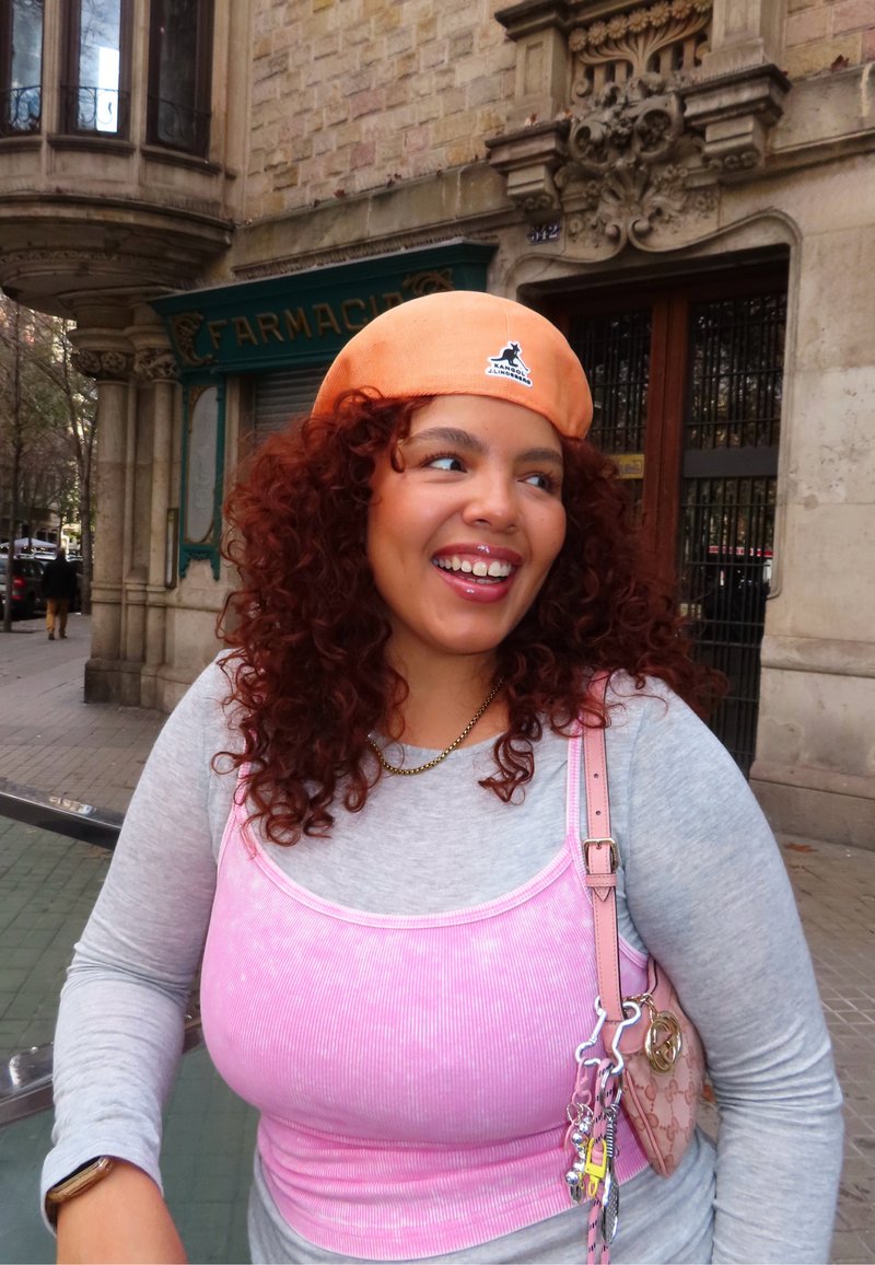 Jeune femme aux cheveux roux bouclés portant une casquette orange, un haut rose sur un t-shirt gris, et portant un sac rose à l'extérieur près d'une vitrine de pharmacie.