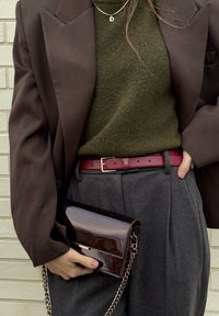 Personne portant un blazer marron, un pull olive, un pantalon gris avec ceinture rouge, tenant une pochette en cuir verni marron foncé avec une bandoulière chaîne dorée.