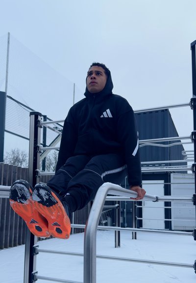 Joven con ropa deportiva negra realizando ejercicio de L-sit en barras paralelas en un área de entrenamiento al aire libre nevada.