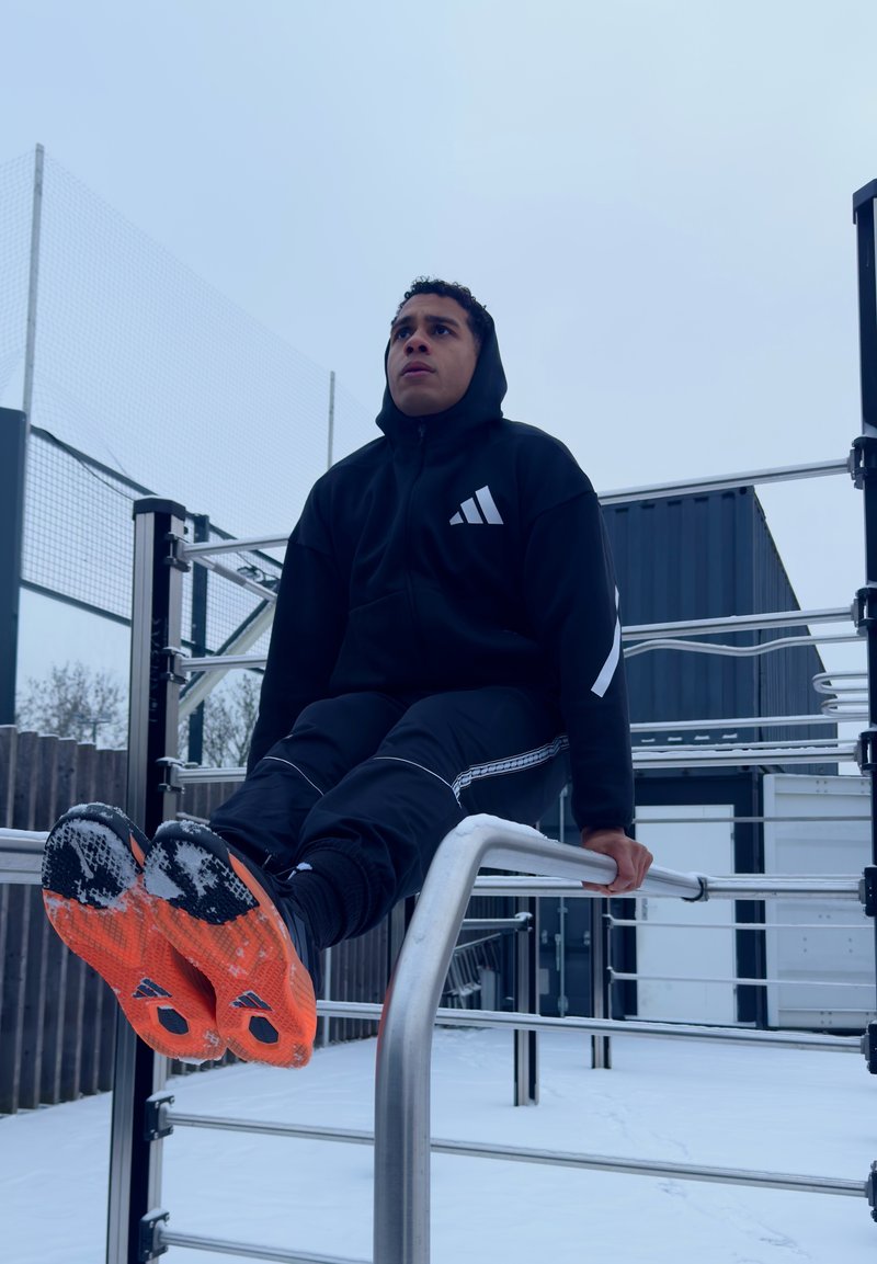 Młody mężczyzna w czarnym stroju sportowym wykonujący ćwiczenie L-sit na poręczach równoległych na zaśnieżonym, zewnętrznym placu do ćwiczeń.