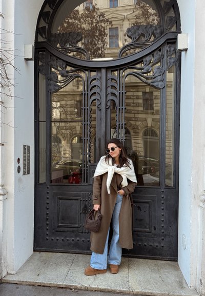Mujer con abrigo marrón, vaqueros azul claro y gafas de sol, de pie frente a unas puertas dobles ornamentadas de metal negro y vidrio.