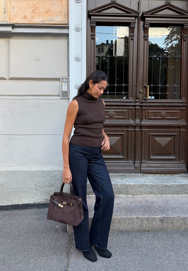 Mujer con jersey de cuello alto sin mangas de color marrón y vaqueros oscuros, sosteniendo un bolso marrón, de pie junto a unas puertas dobles de madera con paneles de vidrio.