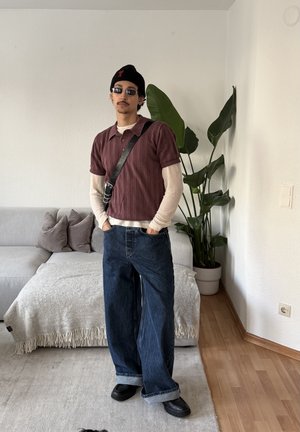 Hombre con varias capas de ropa, pantalones vaqueros anchos, zapatos negros, gorro y gafas de sol, de pie en una sala cerca de un sofá y una planta en maceta.