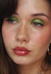 Nahaufnahme eines Gesichts mit grünem Augen-Makeup, einem goldenen herzförmigen Septumring und glänzenden roséfarbenen Lippen, mit Sommersprossen und einem glatten Teint.