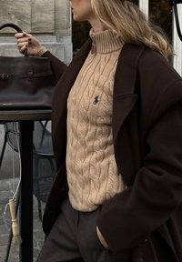 Beige kabelstrikket sweater med høj hals, lagdelt under en mørkebrun overfrakke. Brun håndtaske i nærheden tilføjer tekstur og kontrast.