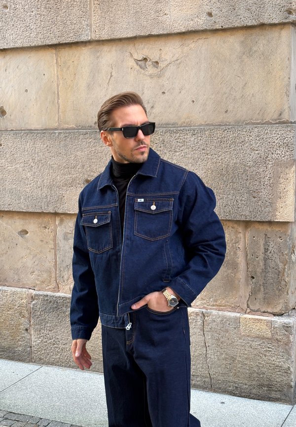 Hombre con chaqueta y pantalones vaqueros azul oscuro, gafas de sol negras y reloj de oro, de pie frente a un fondo de muro de piedra.