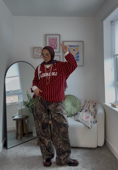 Sudadera de gran tamaño a rayas rojas con el texto "LOS ÁNGELES", pantalones camuflados holgados y zapatos negros de suela gruesa. Los accesorios incluyen gafas y collares.