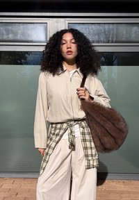 Woman with curly hair wearing a light jacket and wide-leg trousers, with a plaid shirt tied at her waist and holding a large brown furry bag.