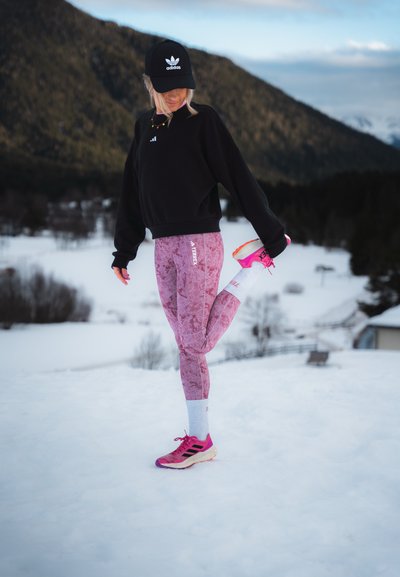Sudadera negra, leggins rosa con estampado, calcetines blancos y zapatillas deportivas de un rosa brillante. Paisaje nevado de fondo con montañas.