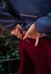 Bordeauxrode sportshorts met de tekst "AEROREADY", gecombineerd met een navyblauw, gestructureerd shirt met lange mouwen. Op de achtergrond is groene bladgroei te zien.