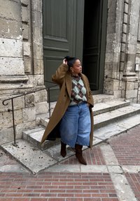 Manteau en laine marron, pull à losanges vert et marron, short en denim bleu et bottes à talons marron. Marches en pierre grise et pavé en briques texturé.