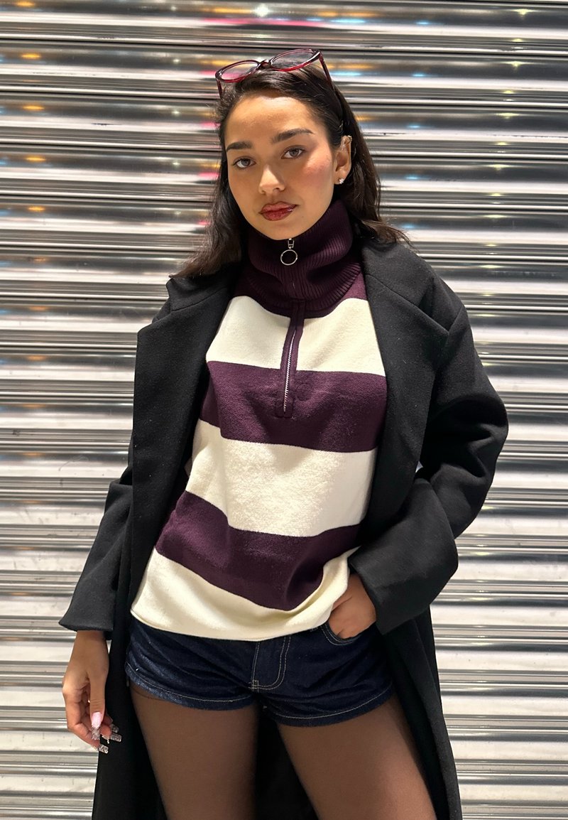 Maglia con mezza cerniera in pile color crema con ampie strisce viola, indossata sotto un cappotto nero. Shorts in denim scuro completano l'outfit.