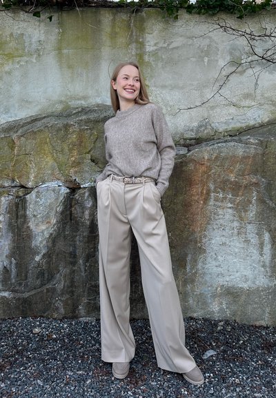 Mujer joven sonriendo, vestida con suéter beige y pantalones anchos plisados, de pie sobre grava frente a un fondo de roca texturizada y pared.