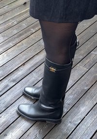 Zwarte lederen kniehoge laarzen met platte zolen, voorzien van een ritssluiting aan de zijkant en decoratieve gouden accenten. Gedragen over zwarte panty's.