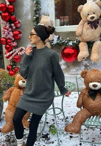 Femme en pull gris et leggings noirs se tient près d'ornements de Noël rouges et de grands ours en peluche avec de la fausse neige en plein air.