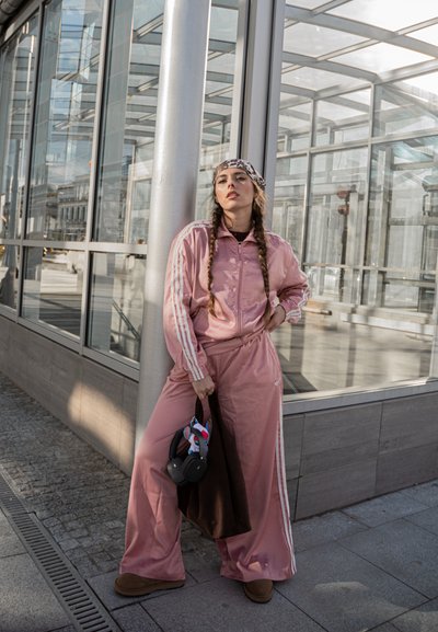 Chándal rosa con acentos de rayas blancas, pantalones anchos y una chaqueta con cremallera. Se puede ver un pañuelo estampado y un bolso marrón con un acento de juguete.