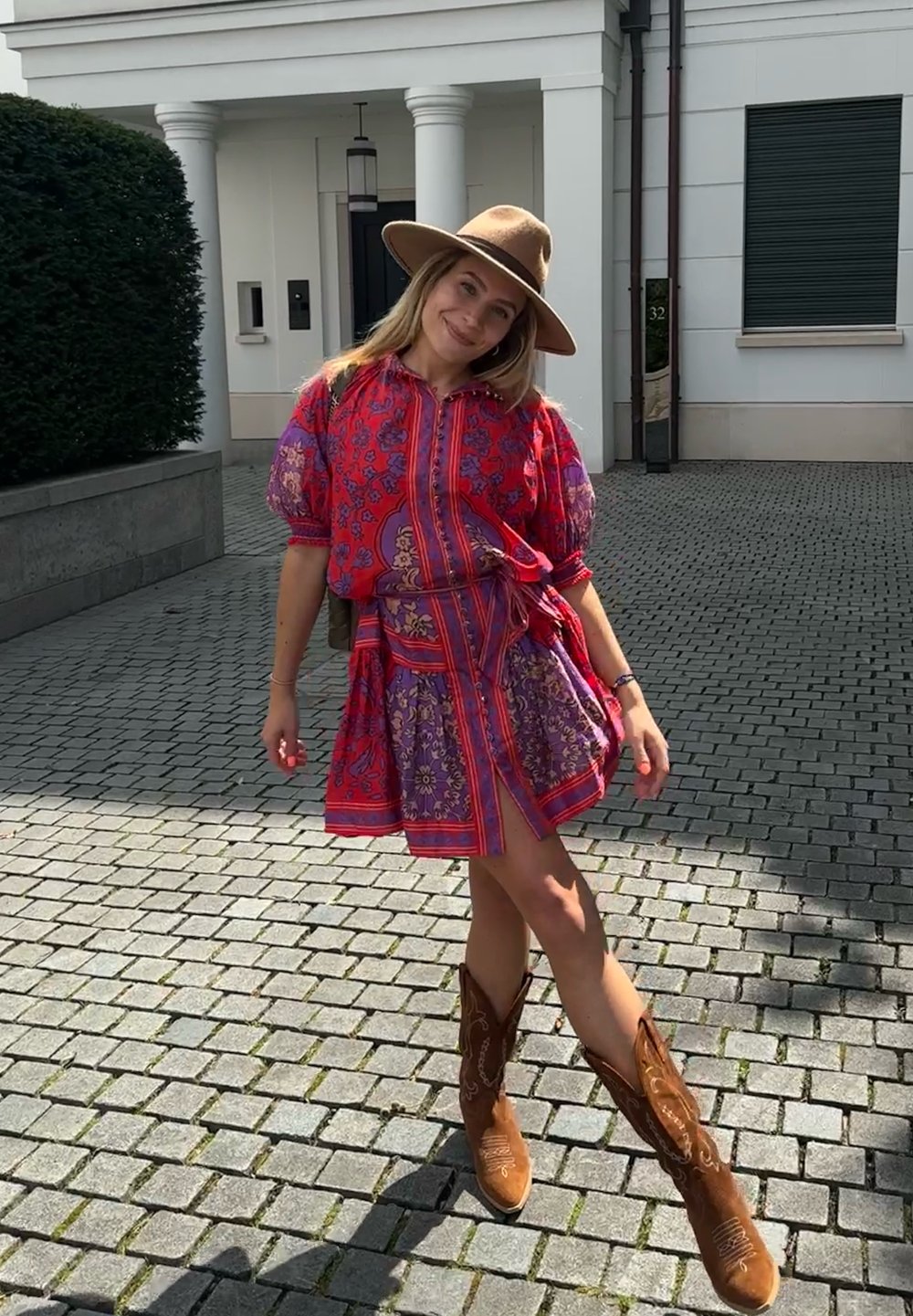 Rochie scurtă colorată cu modele florale și legături, asortată cu cizme cowboy maro. În aer liber, pe paviment din piatră, cu o casă în fundal.