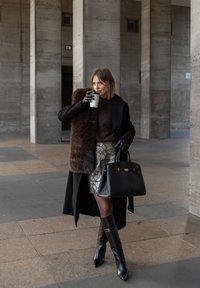 Čierny kabát s kožušinovou lemovkou, hnedý sveter, sukňa s hadím vzorom, čierne čižmy po kolená a čierna kabelka. Drží pohár na kávu so sebou.