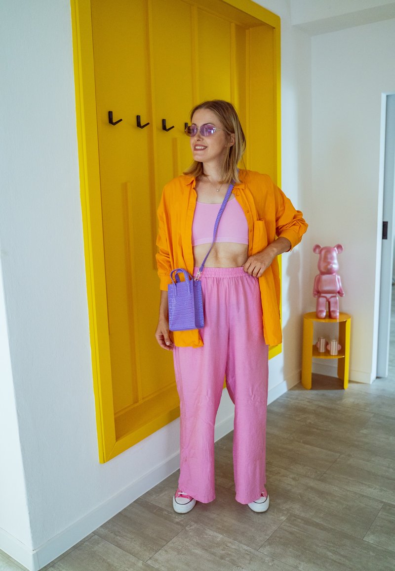 Camisa laranja brilhante com botões, top cropped cor de rosa e calças soltas cor de rosa. Pequena bolsa roxa com acabamento texturizado. Parede amarela com ganchos atrás.