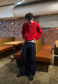 Pull-over rouge en tricot avec un col et trois boutons, associé à un jean large bleu foncé. Debout dans un cadre intérieur décontracté.