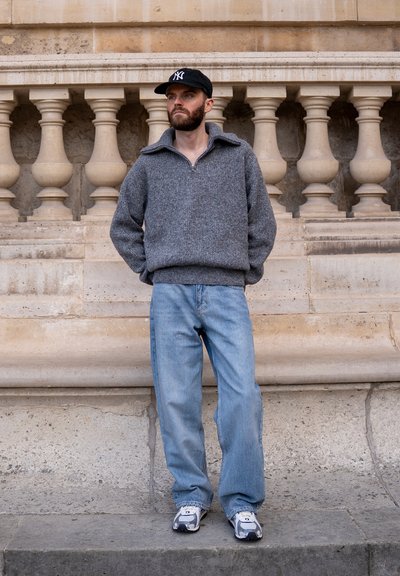 Hombre con suéter gris, vaqueros azul claro, zapatillas deportivas y gorra negra, de pie con las manos en los bolsillos junto a una balaustrada de piedra.
