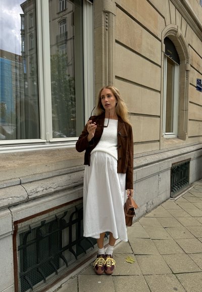Chaqueta de ante marrón sobre un suéter blanco de canalé y una falda maxi blanca fluida. Bolso marrón y zapatillas multicolores con cordones amarillos.