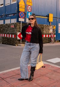 Jachetă din denim negru, tricou grafic roșu Puma, blugi largi bleu deschis, cizme negre până la genunchi și o geantă mică galbenă. Fundal urban.