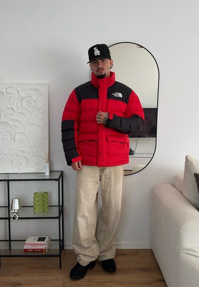 Chaqueta acolchada en rojo y negro con un cuello alto, que presenta un logo y dos bolsillos frontales. Llevada sobre pantalones sueltos beige y zapatos negros.